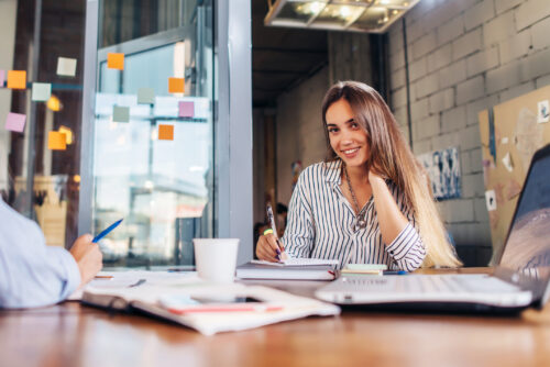 Portrait Of Smiling Female Office Worker Writing Looking At Camera Sitting At Conference Room During The Meeting